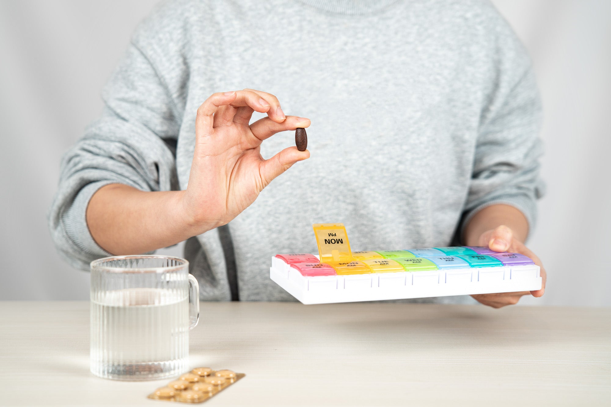 a person in a gray sweatshirt holds a pill in one hand and a rainbow pill case in the other. There is a glass of water by them